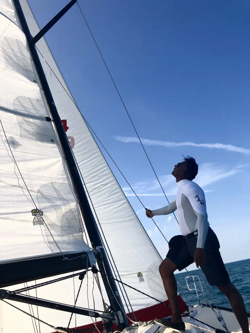 Dhamitha Arsakularatna at the mast of a racing sailboat, one hand on a stay, looking up at the sails against a blue sky